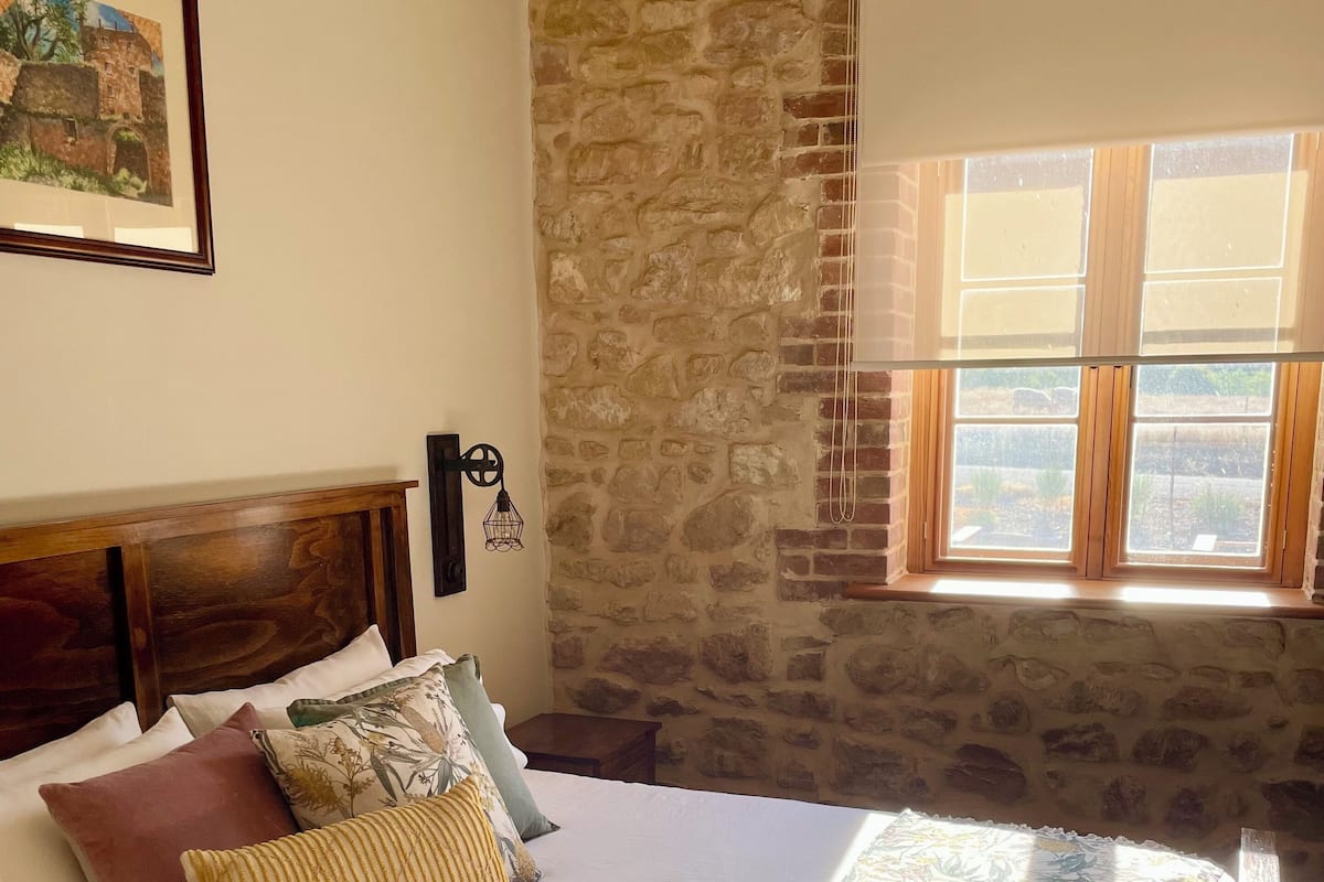 Un dormitorio con pared de piedra, cabecero de madera, cama con ropa de cama blanca y varias almohadas, un plaid decorativo y una ventana con vistas a la vegetación.