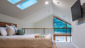 Une chambre à coucher avec un lit, des fenêtres de toit, une télévision et un balcon donnant sur la forêt.