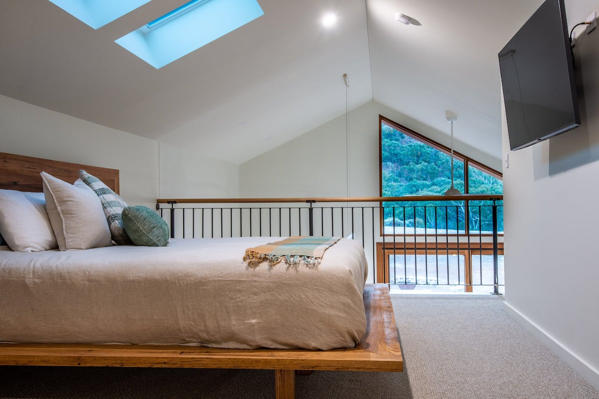 Un dormitorio con cama, tragaluces, televisor y balcón con vista a árboles.