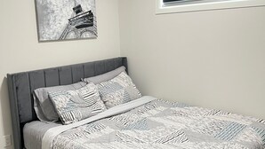 A bedroom with a bed, a gray headboard, a ceiling light, a window with blinds, and a wall-mounted picture of the Eiffel Tower.