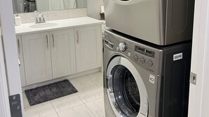 A modern laundry room with stacked front-loading washers and dryers, a white tiled floor, and a large mirror above the sink.