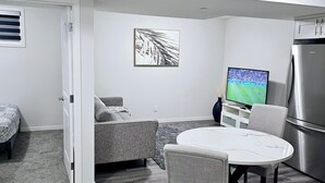 A modern living room with a grey sofa, a round dining table with chairs, a flat-screen TV, and a stainless steel refrigerator.