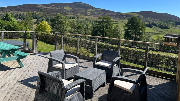 A deck with wicker furniture and a table, overlooking a scenic landscape with hills and trees.