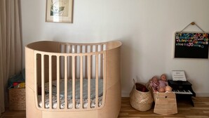 Una cuna para bebé con acabado de madera, un cuadro en la pared, un pequeño piano y una cesta con juguetes.