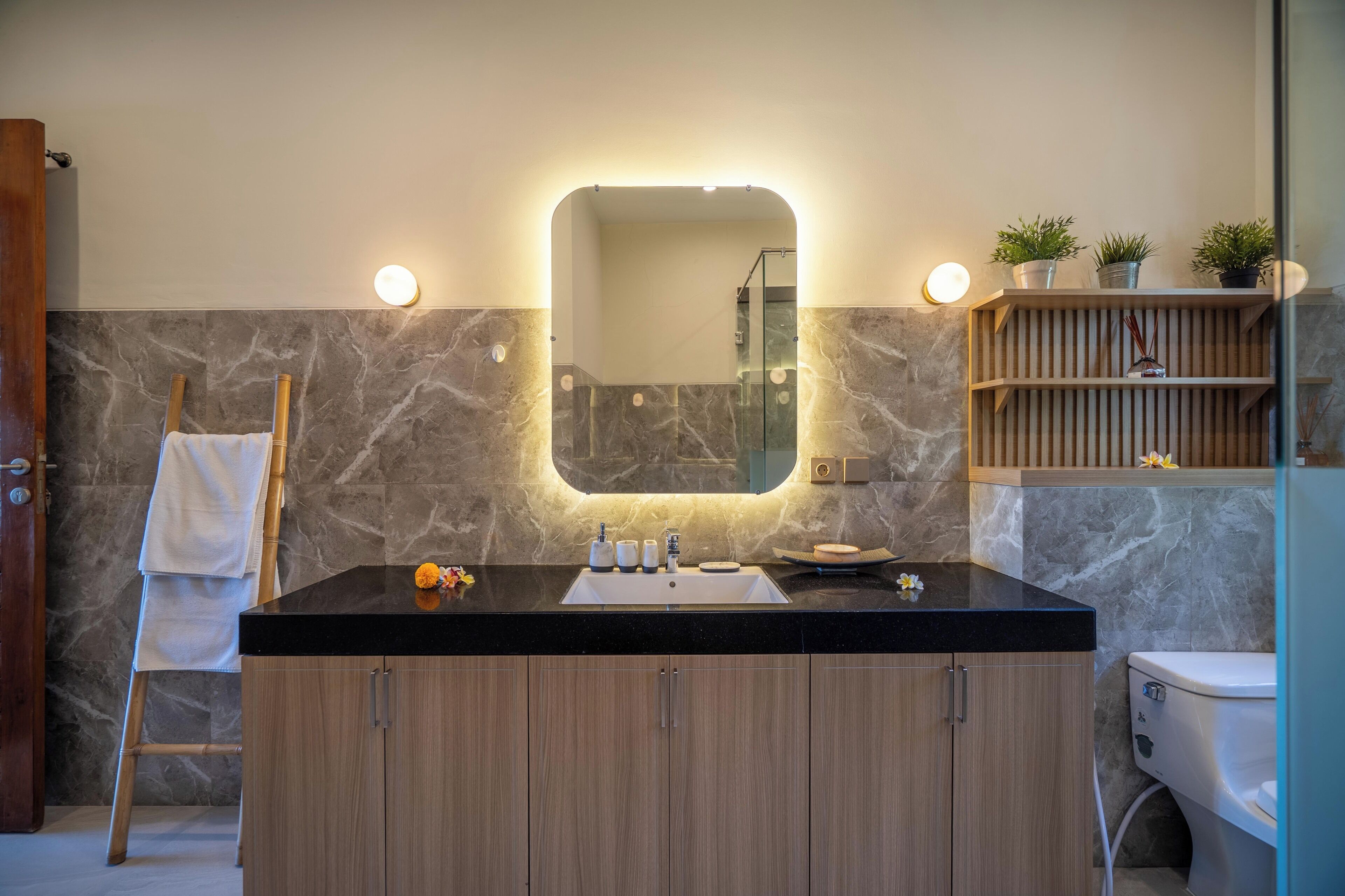 A modern bathroom with a marble wall, a wooden vanity, a black countertop, and a mirror with lights.