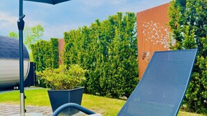 A sunlit outdoor seating area with a lounge chair and a blue umbrella, surrounded by greenery and a decorative wall.