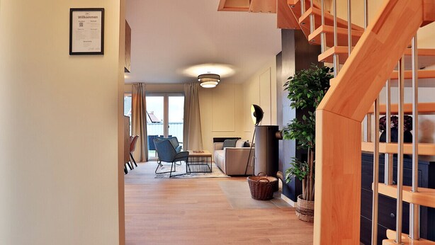 A spacious living area with a wooden staircase, a sofa, a dining table, and a large window.