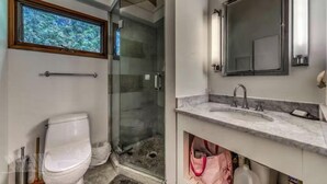 Une salle de bain comprenant des toilettes, une douche fermée par une paroi de verre, un lavabo avec un miroir et une fenêtre donnant sur les arbres.