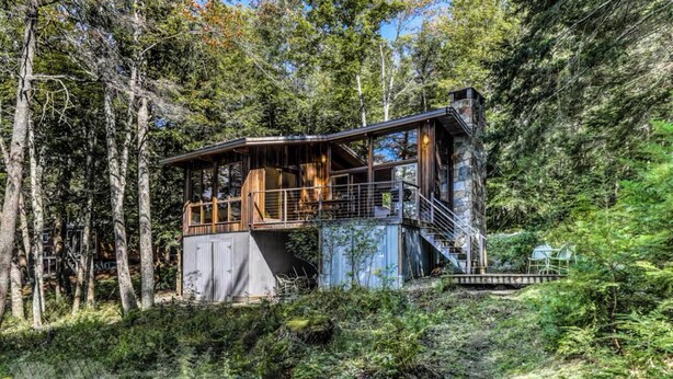 Une cabane dotée d’une terrasse en bois et d’une cheminée en pierre, entourée d’une forêt dense.