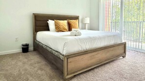 Een slaapkamer met een groot bed, een nachtkastje, een lamp en uitzicht op de omgeving door een raam met jaloezieën.