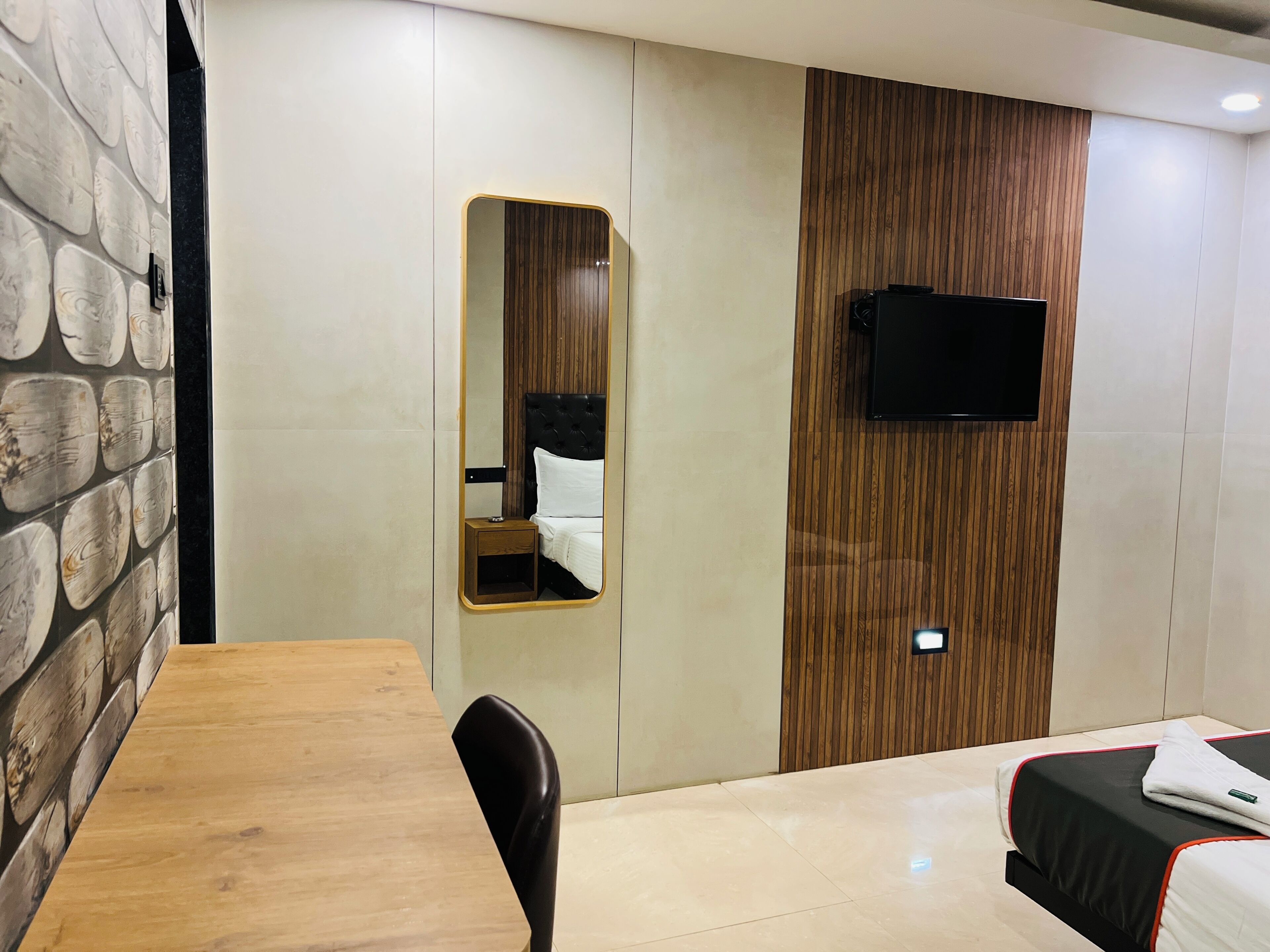 A hotel room with a wooden dining table, a single black chair, a bed with a black and white bedspread, a wooden panel wall, and a mounted television.