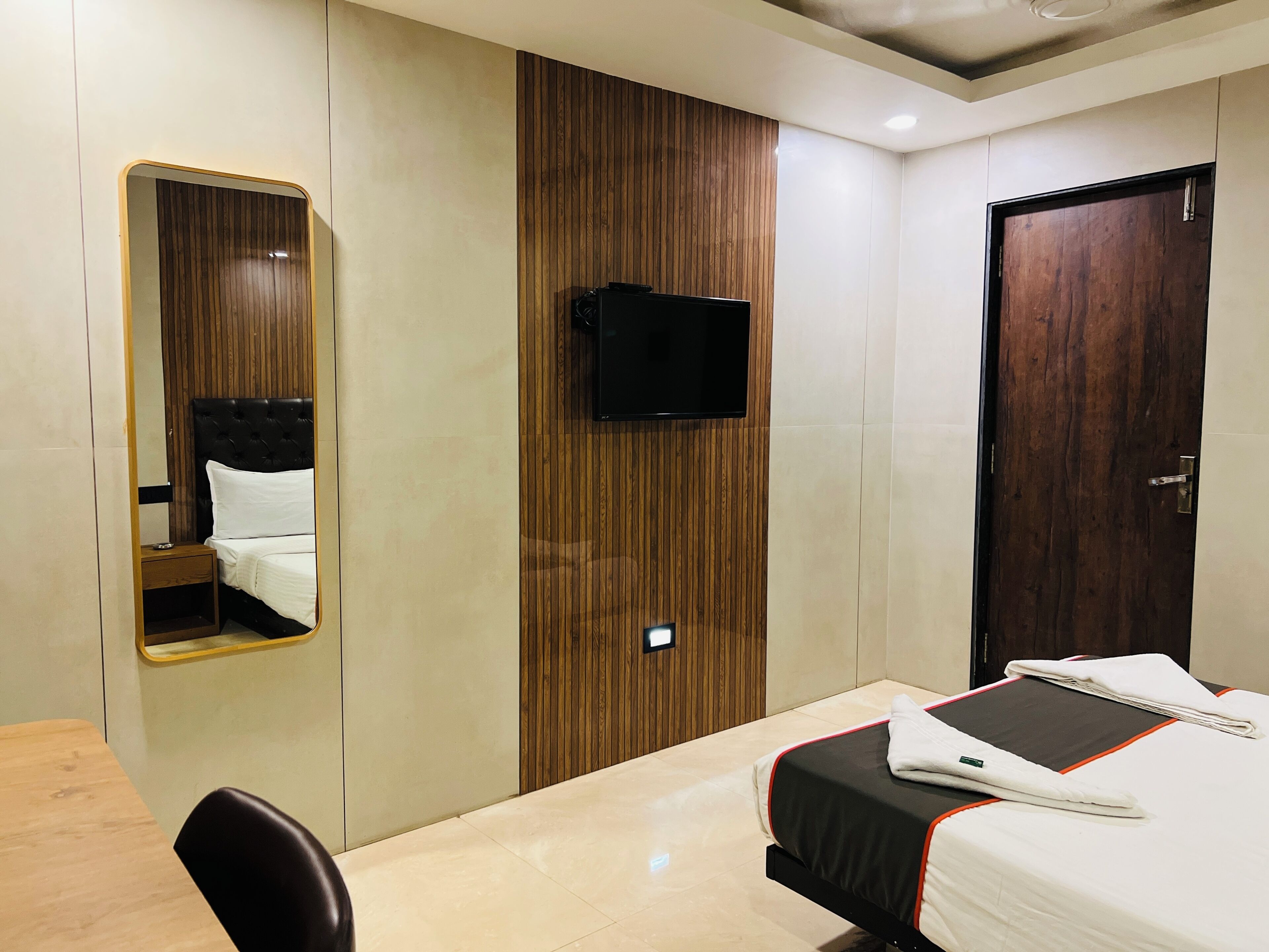 A hotel room with a bed, a TV mounted on the wall, a wooden wardrobe, a mirror, and a door.