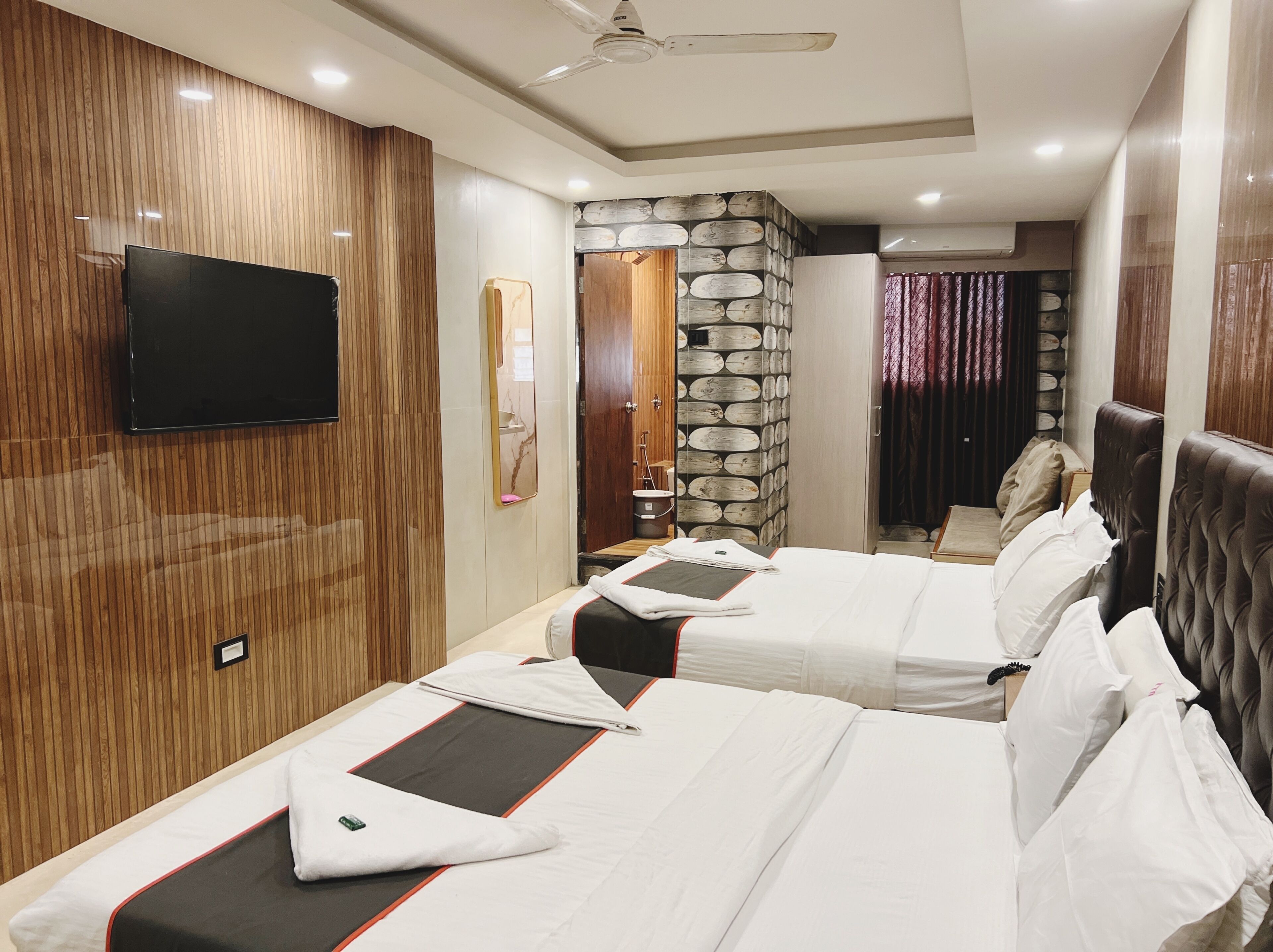 A hotel room with two beds, a flat-screen TV, a wooden panel wall, a stone accent wall, and a ceiling fan.