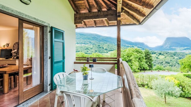 Een balkon met een glazen tafel en stoelen, een houten plafond en uitzicht op een groene vallei en bergen.