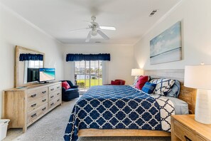 Une chambre à coucher comprenant un lit, une commode, un téléviseur, un ventilateur de plafond, une fenêtre avec des rideaux et un tableau accroché au mur.