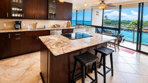 A modern kitchen with dark wood cabinets, a marble countertop, and a large island with stools. - Renee's Poipu Paradise! Oceanfront Views & A/C! (Koloa)