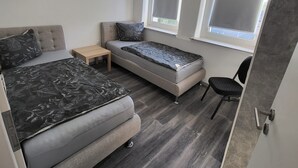 Une chambre à coucher de taille modeste, comprenant un lit, un petit canapé et une chaise. Elle est dotée de deux fenêtres équipées de stores et le sol est en bois.
