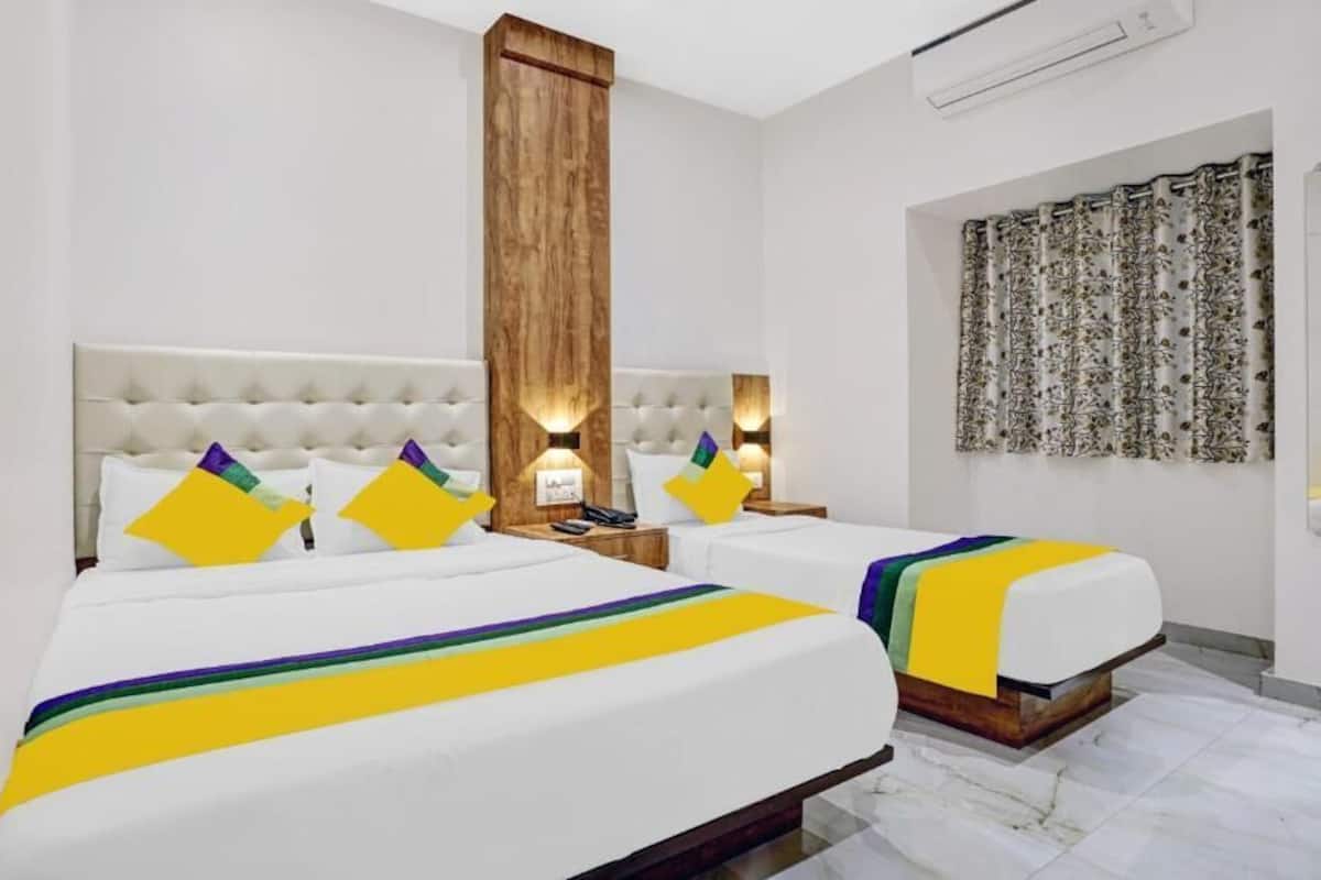 Habitación de hotel con dos camas, cada una con cabecero blanco y un panel de pared de madera. Hay dos mesitas de noche con lámparas, y una cortina con estampado floral en la ventana.