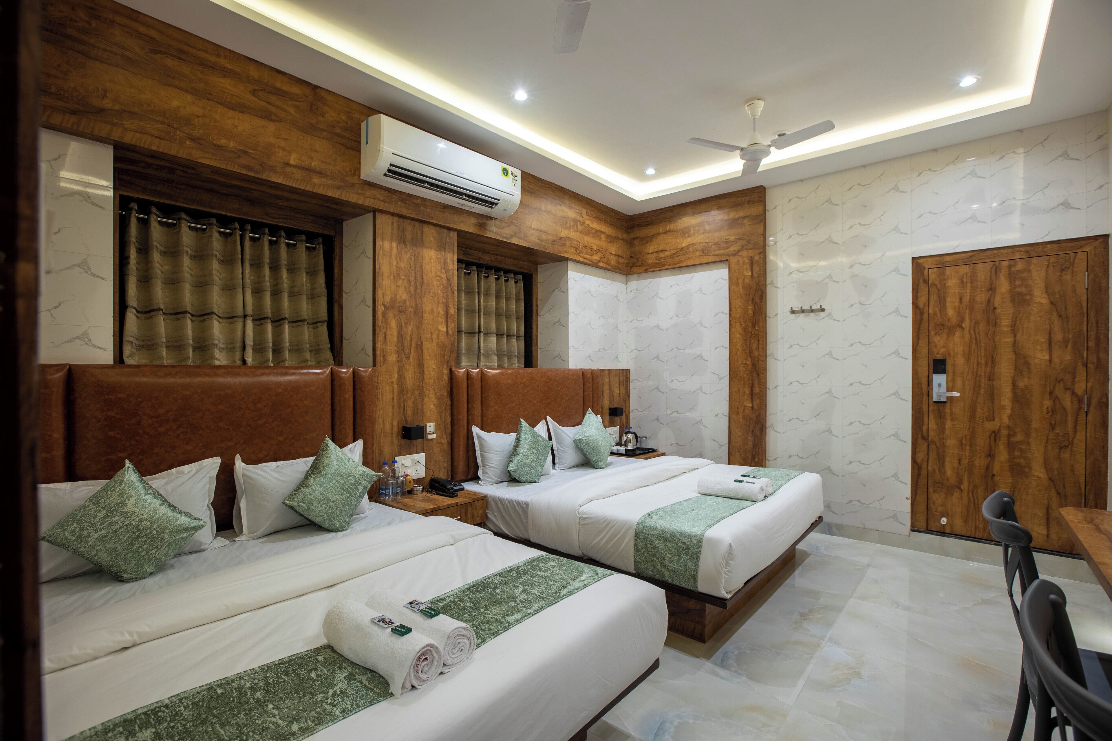 Een hotelkamer met drie bedden, houten hoofdborden, een plafondventilator, een airconditioning en een houten garderobe.