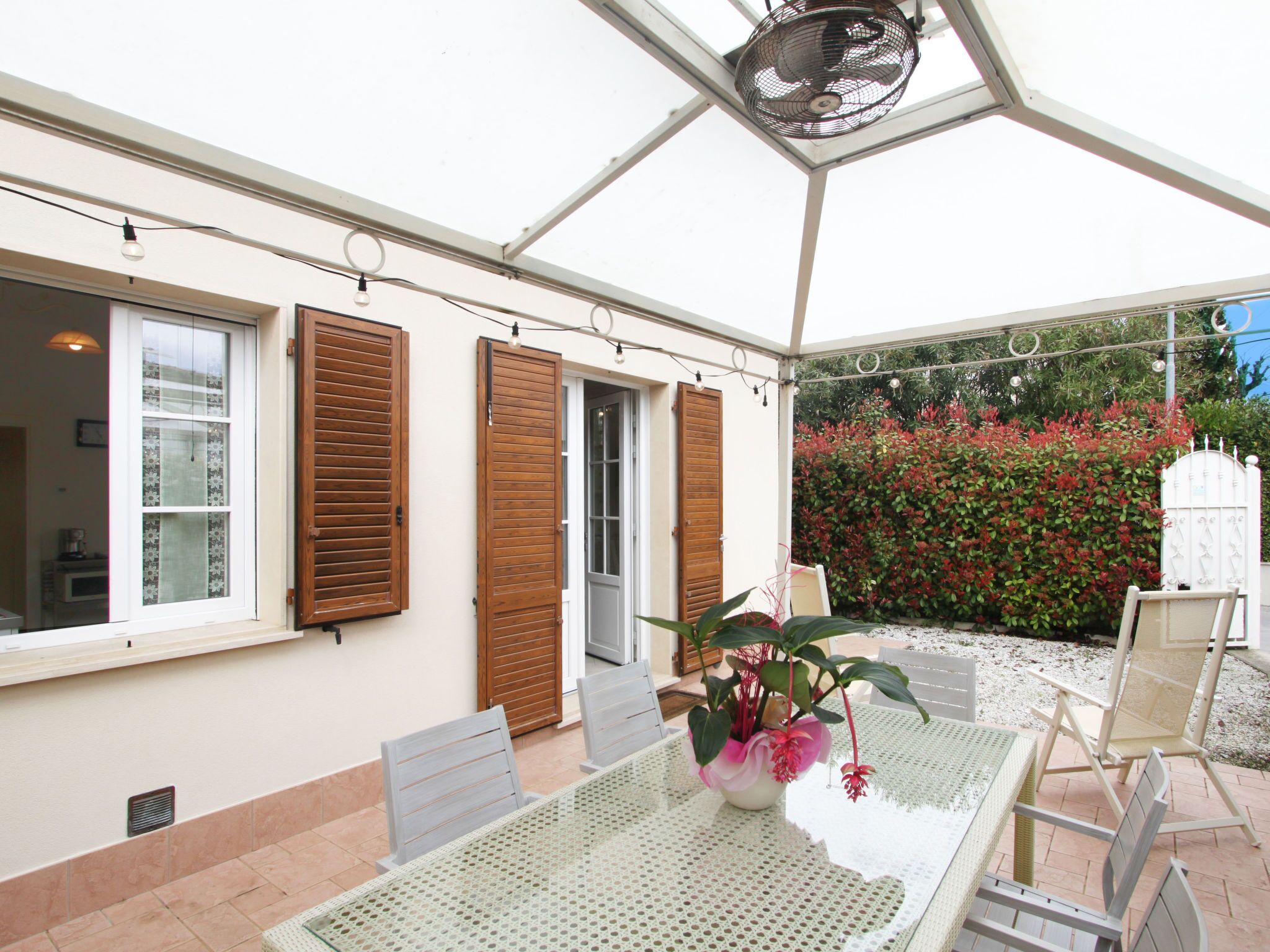 A covered patio with a table and chairs, a potted plant, and a view of a garden with red foliage.