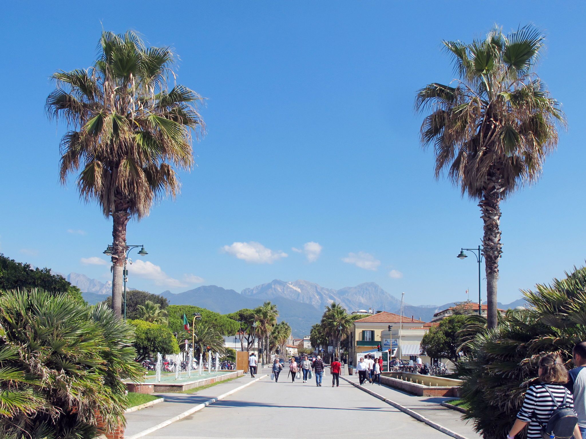 Une large promenade bordée de palmiers et agrémentée d’une fontaine, qui mène à un point de vue sur les montagnes.