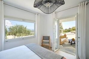Ein Schlafzimmer mit einem großen Bett, einem Sessel und Blick auf eine Terrasse mit Tisch und Kissen.
