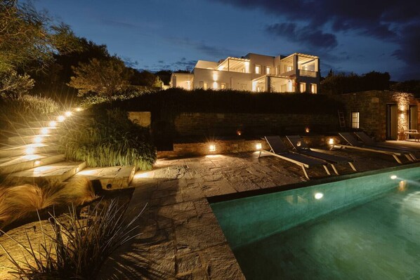 A modern house with a swimming pool, outdoor seating, and a well-lit garden at night.