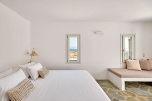 A bedroom with a large bed, a bench, and two windows offering a view of the landscape.