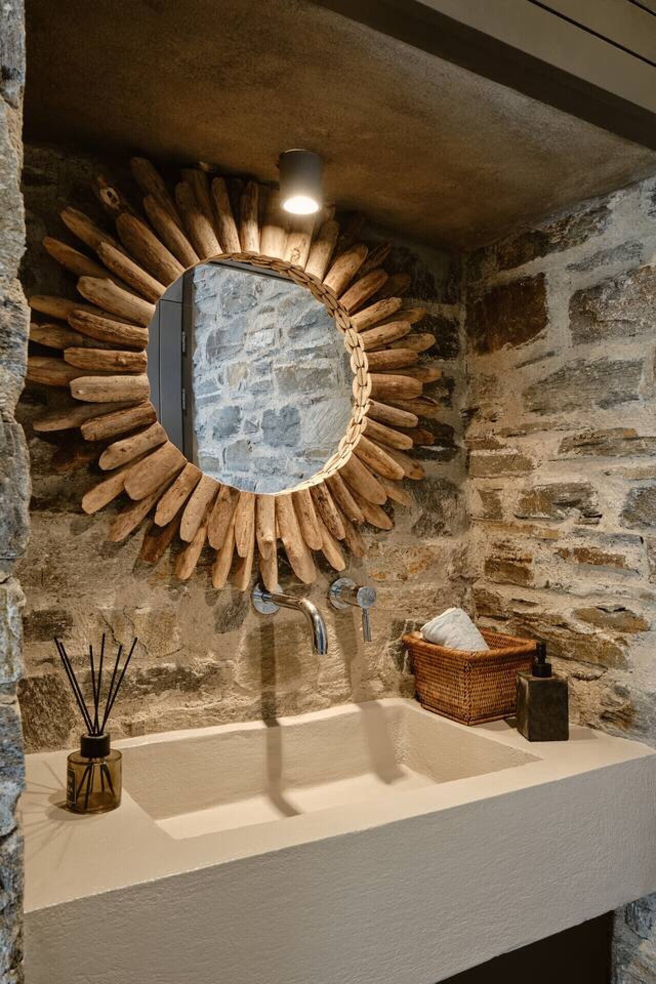Un baño con pared de piedra, espejo con marco de madera, lavabo y una cesta.