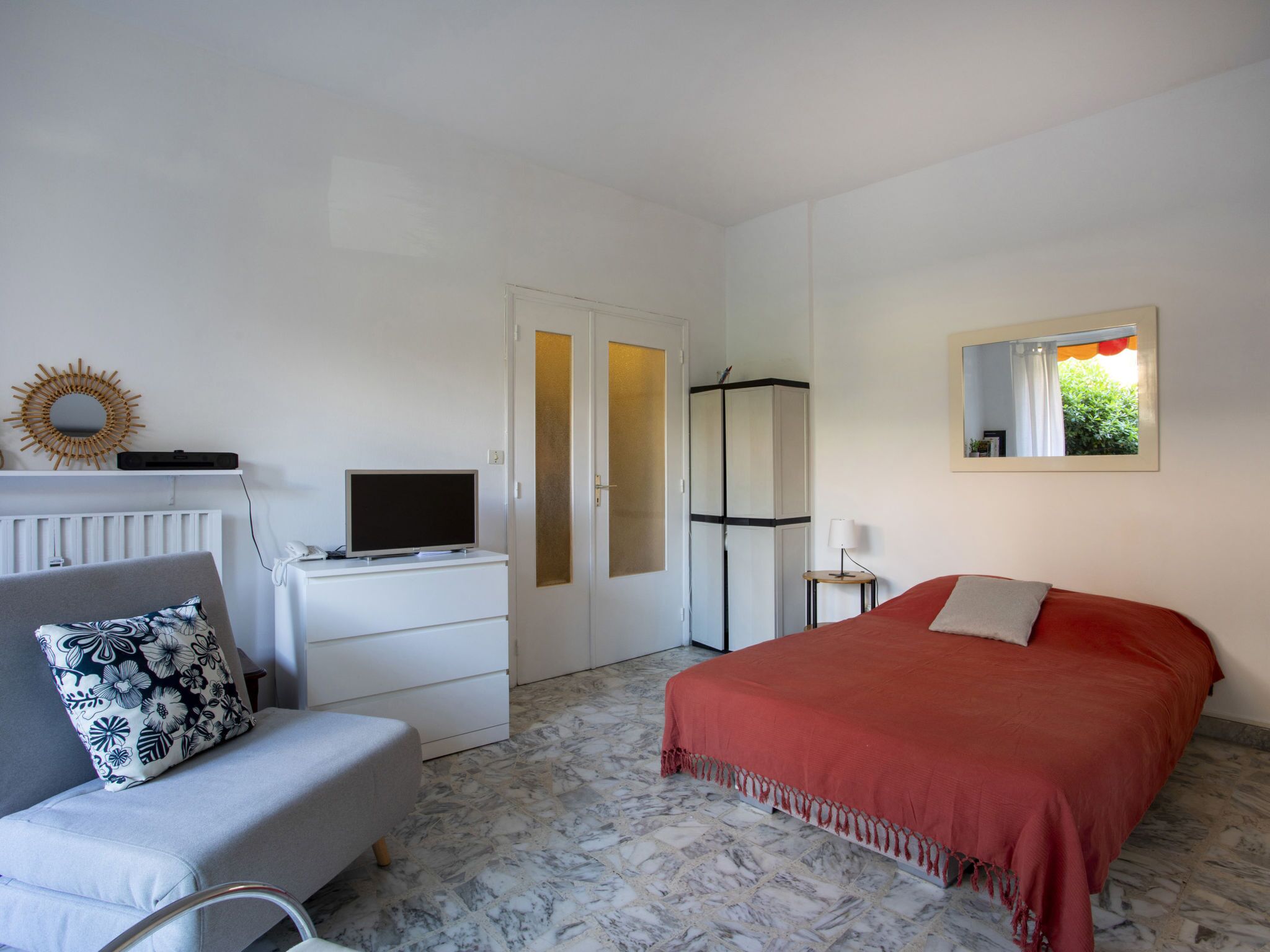 Ein Schlafzimmer mit einem Bett, einem Fernseher auf einer Kommode, einem Sessel, einem Kleiderschrank und einem Spiegel an der Wand.