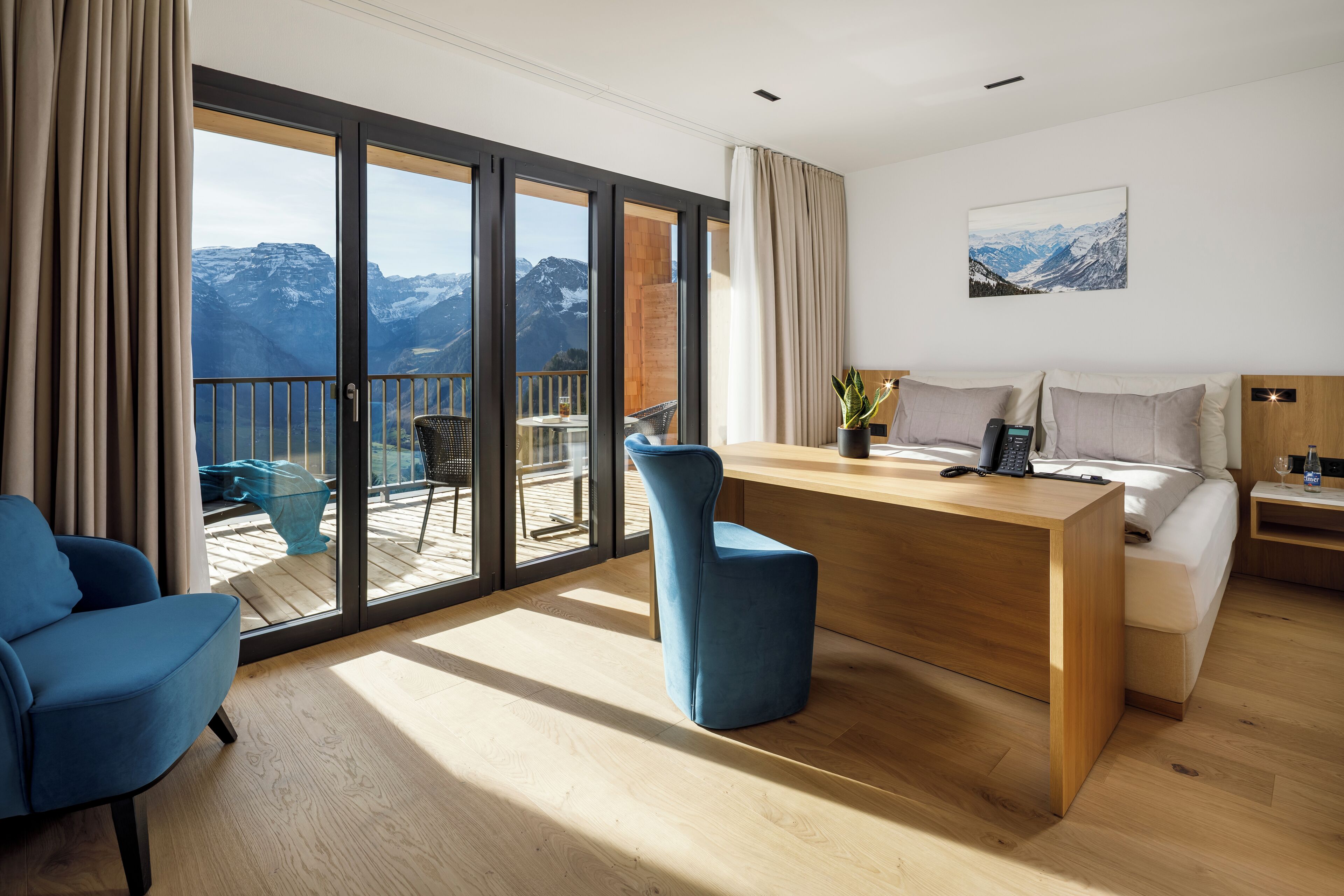 Una habitación de hotel moderna con un escritorio de madera, una cama, un sillón azul y un balcón con vistas a la montaña.