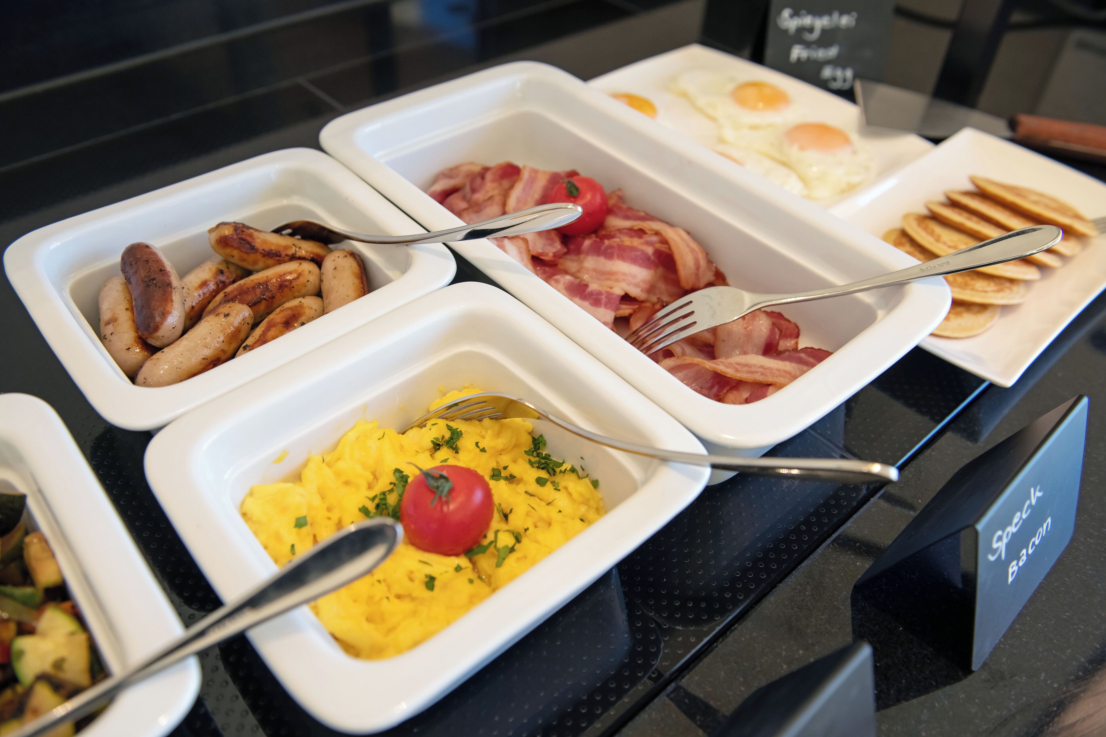 A buffet with various breakfast options including sausages, bacon, eggs, pancakes, and scrambled eggs.