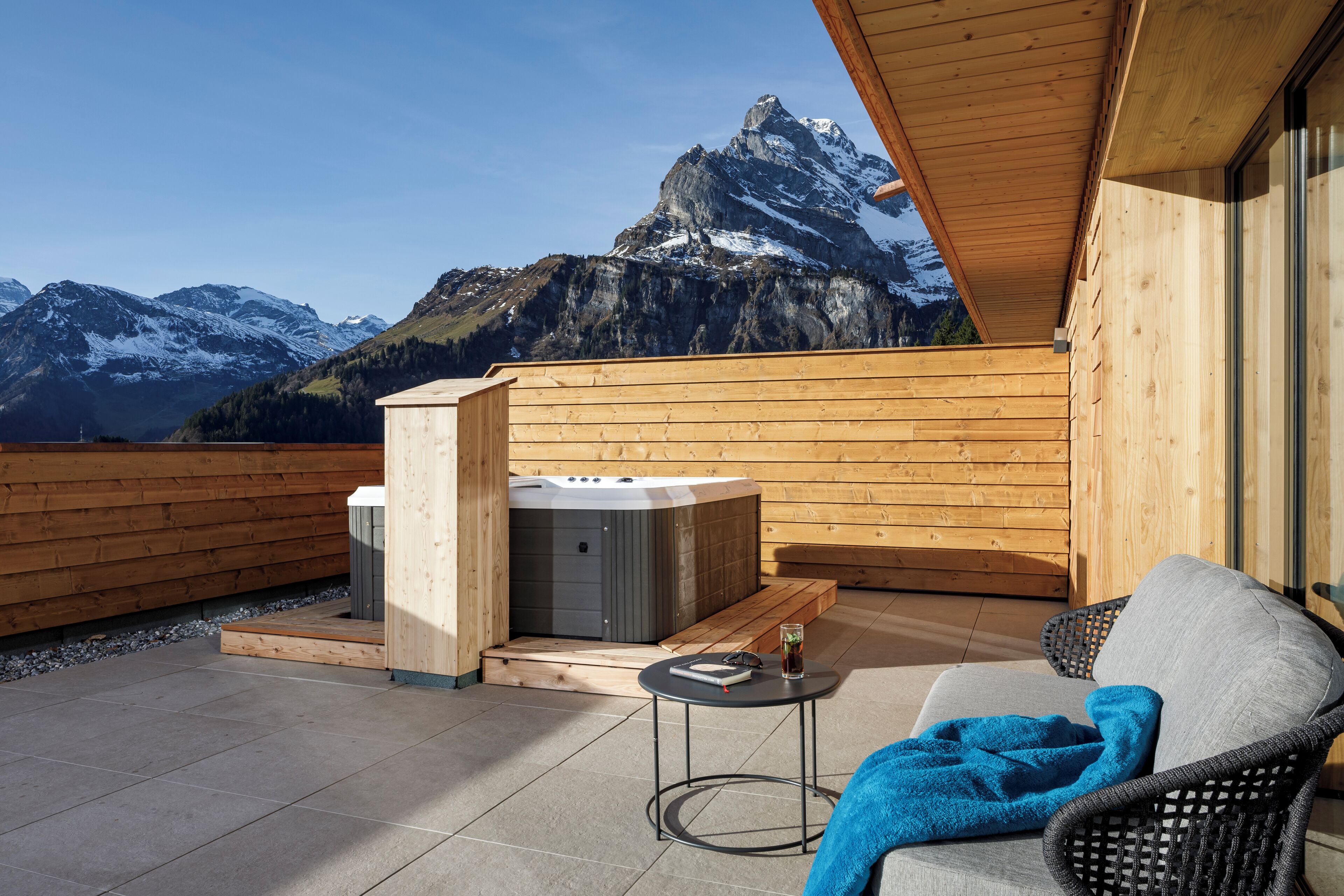 A wooden deck with a hot tub, a table with a drink, and a chair with a blanket.