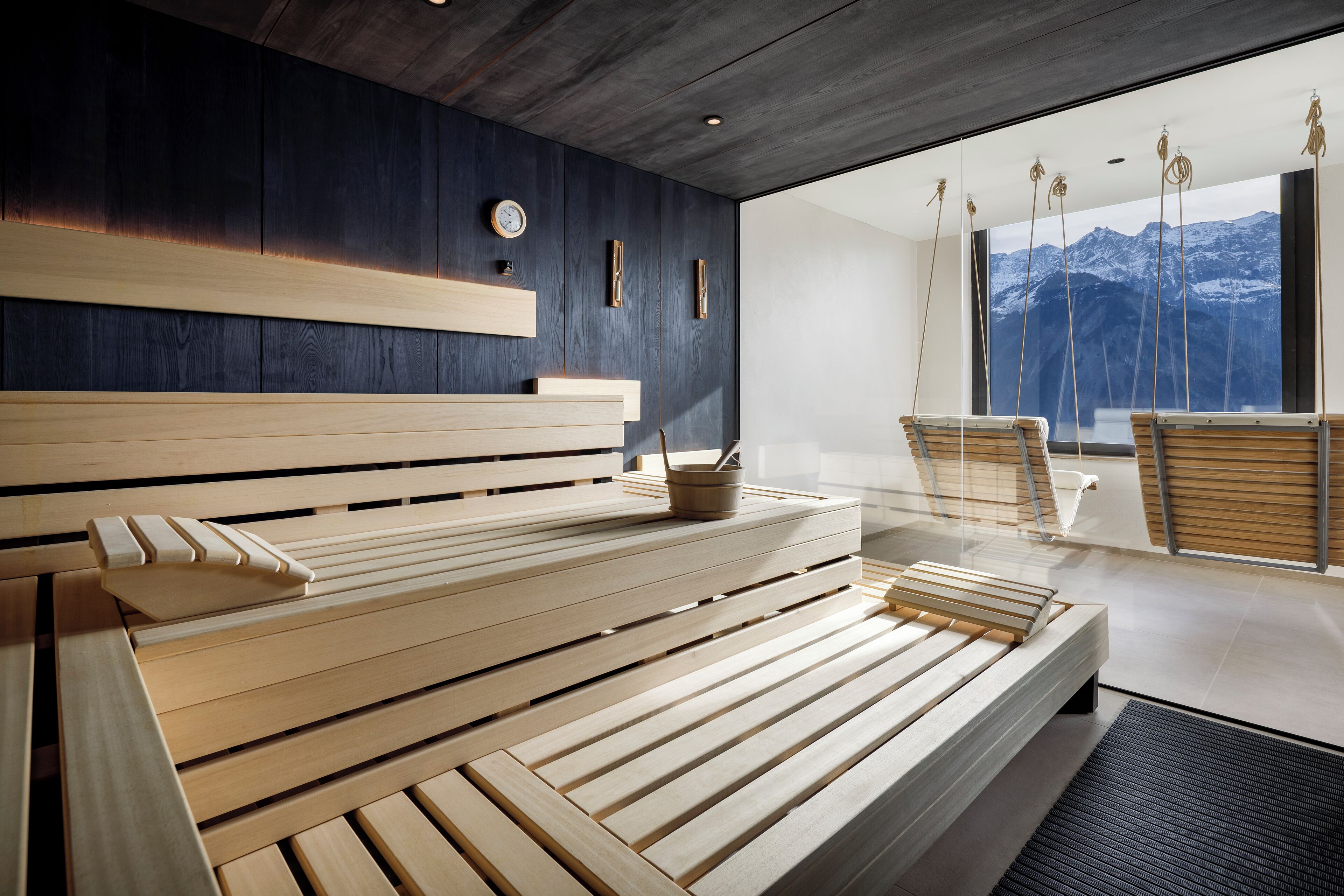 Una moderna sala spa con una panca di legno, un tavolino con una tazza e un cucchiaino, e vista sulle montagne attraverso ampie finestre.