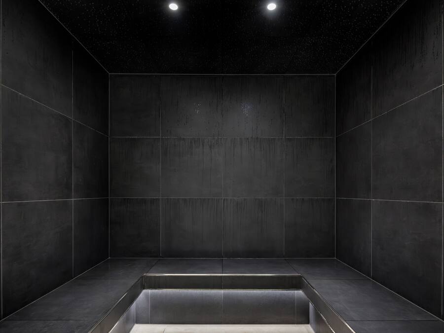 Una habitación con paredes de baldosas oscuras, un banco central y dos luces en el techo.