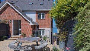 Ein Backsteinhaus mit Terrasse, darauf ein runder Tisch und Stühle, umgeben von Blumentöpfen und einem Holzzaun.