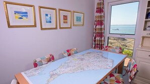 Ein Zimmer mit einer großen Karte auf dem Tisch, bunten Stühlen, einem Fenster mit Meerblick und gerahmten Bildern an der Wand.