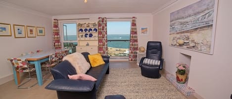 A living room with a sofa, a chair, a dining table, and a view of the sea.