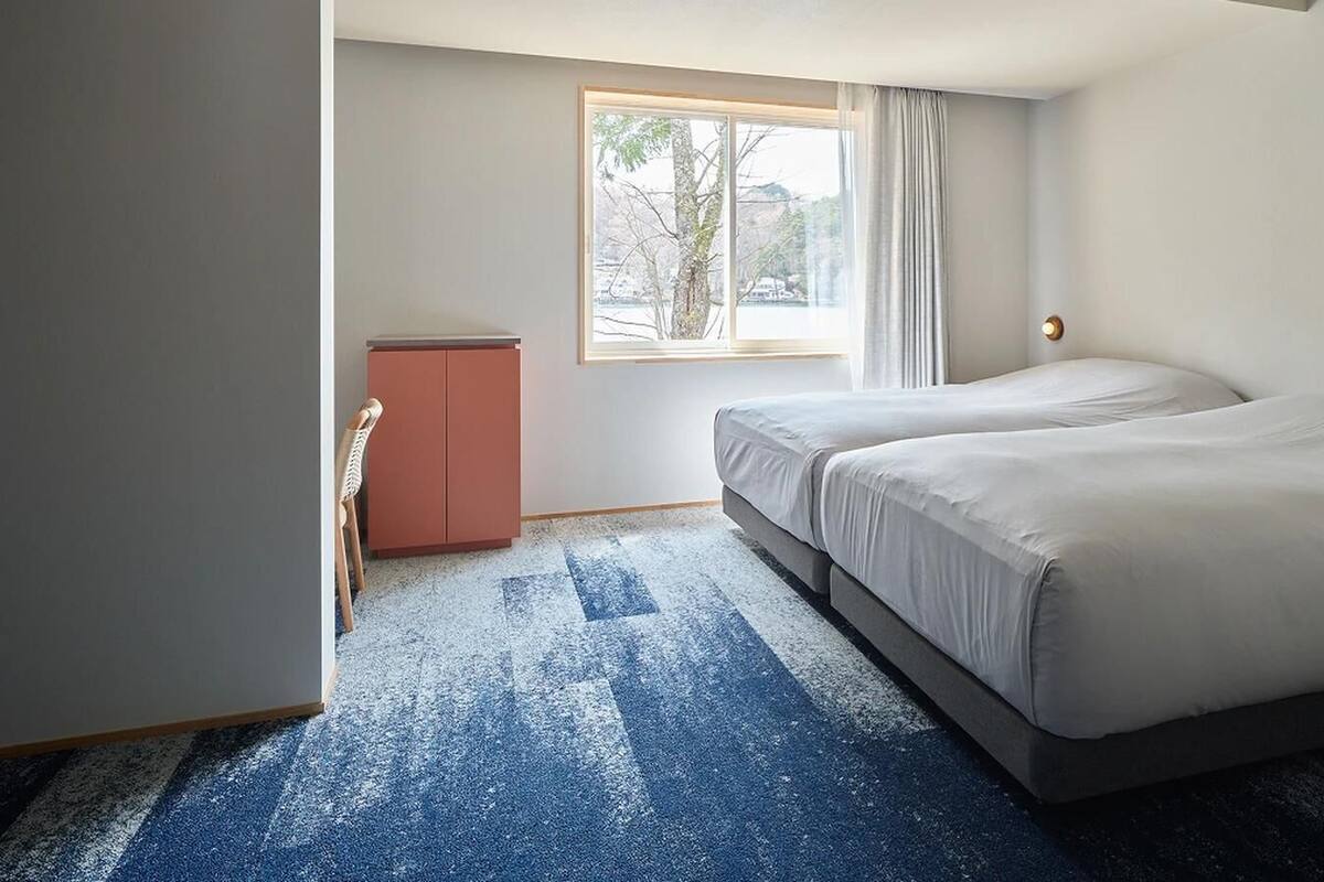 Habitación de hotel con dos camas, un escritorio pequeño y ventana con vista a árboles.
