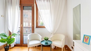 Een kamer met een houten vloer, twee witte stoelen, een klein rond tafeltje met een plant, een grote spiegel en een deur die naar buiten leidt.