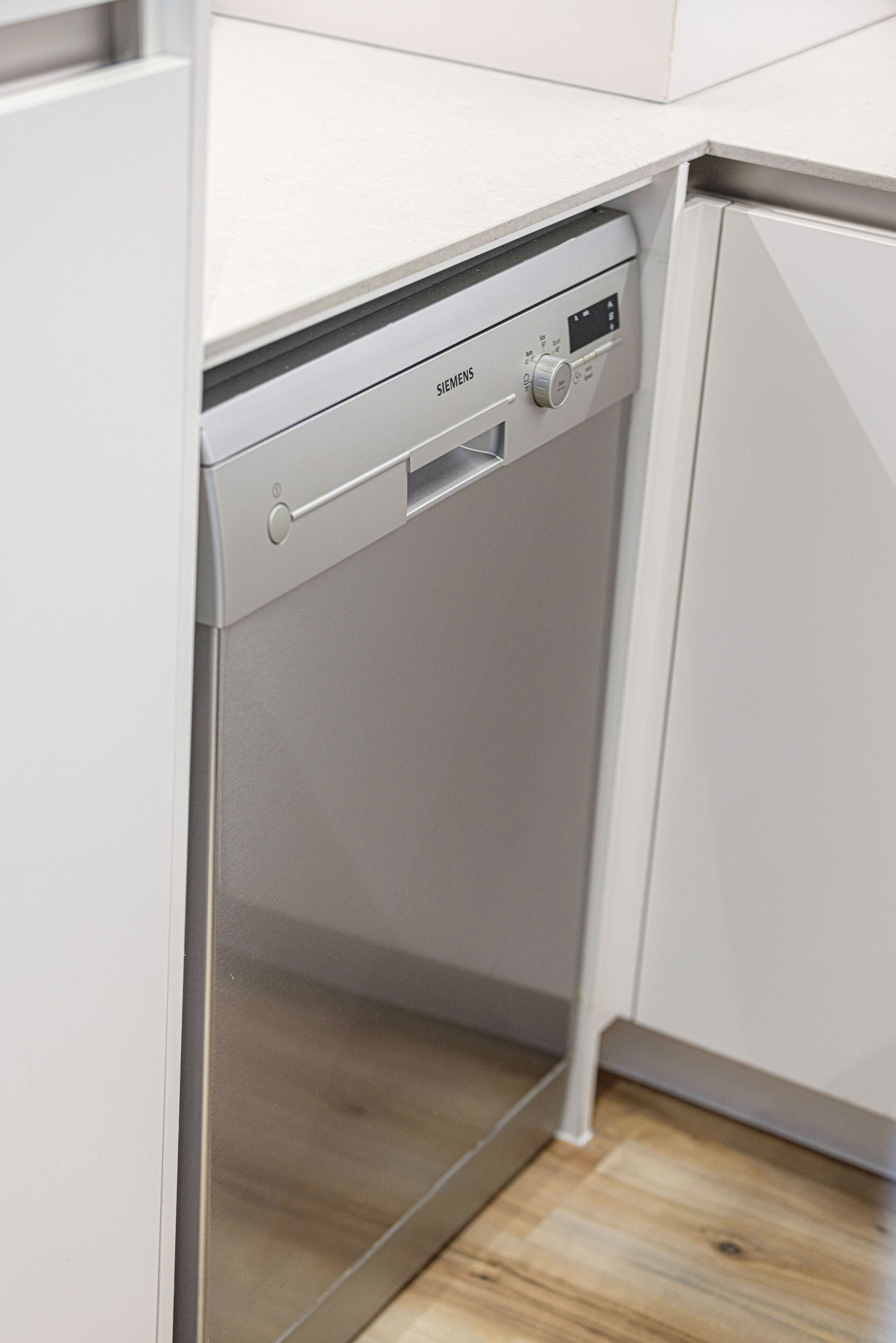 Una cocina compacta y moderna con lavavajillas integrado, muebles blancos y suelo de madera.