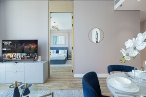 A modern living room with a flat-screen TV displaying a show, a dining area with a table set for two, and a bedroom visible through an open door. - Modern Coastal Luxe  JBR Apartment (Dubai)