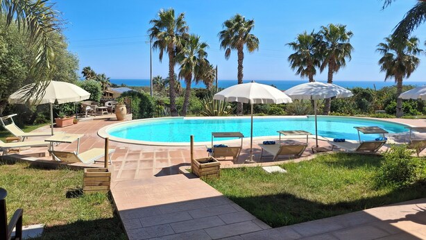 Zona de piscina con sillones de descanso y sombrillas, rodeada de palmeras y vistas al océano.