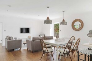 A modern living room with a flat-screen TV, grey sofas, wooden chairs, and pendant lights.