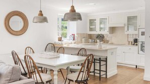 Cocina moderna con armarios blancos, una isla central, iluminación colgante y un comedor con sillas de madera.