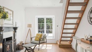 Une pièce avec une cheminée, un escalier en bois, une fenêtre donnant sur la verdure et un fauteuil avec un coussin.