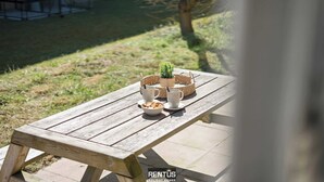 A wooden picnic table with a bowl of cookies and two white cups, set on a patio with a grassy area and a building in the background. - James Ensor Zon - beach fun in Oostduinkerke (Oostduinkerke)