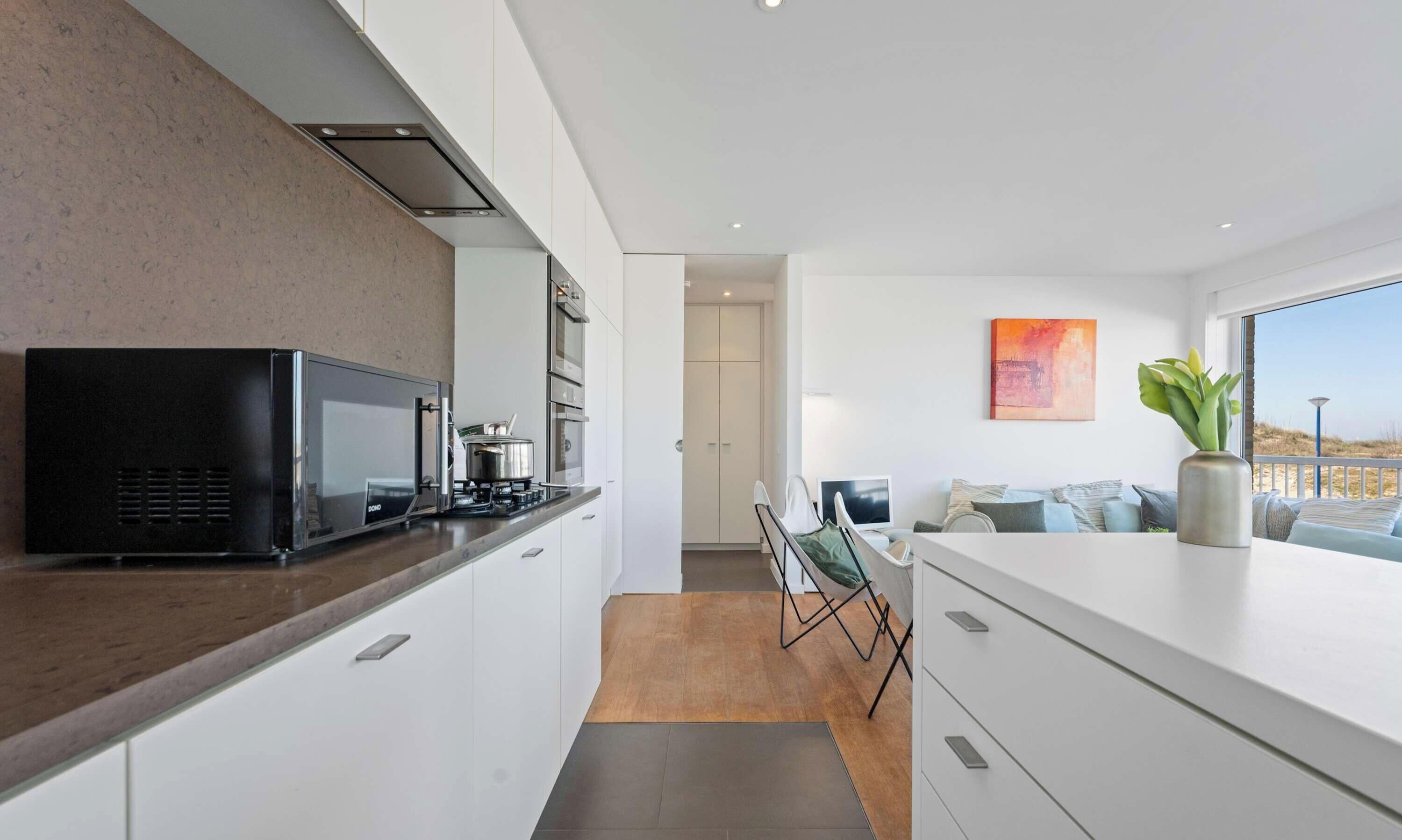 A modern kitchen with a black microwave, stove, and white cabinets. There is a dining area with a table and chairs, and a large window offering a view of the beach.