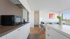 Une cuisine moderne avec un micro-ondes noir, une cuisinière et des armoires blanches. On y trouve un coin repas avec une table et des chaises, ainsi qu’une grande fenêtre donnant sur la plage.