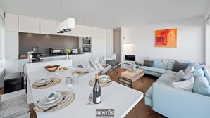 Une cuisine moderne avec des meubles blancs, un coin repas avec une table blanche dressée pour six personnes, et un salon avec un canapé bleu clair et un téléviseur à écran plat.